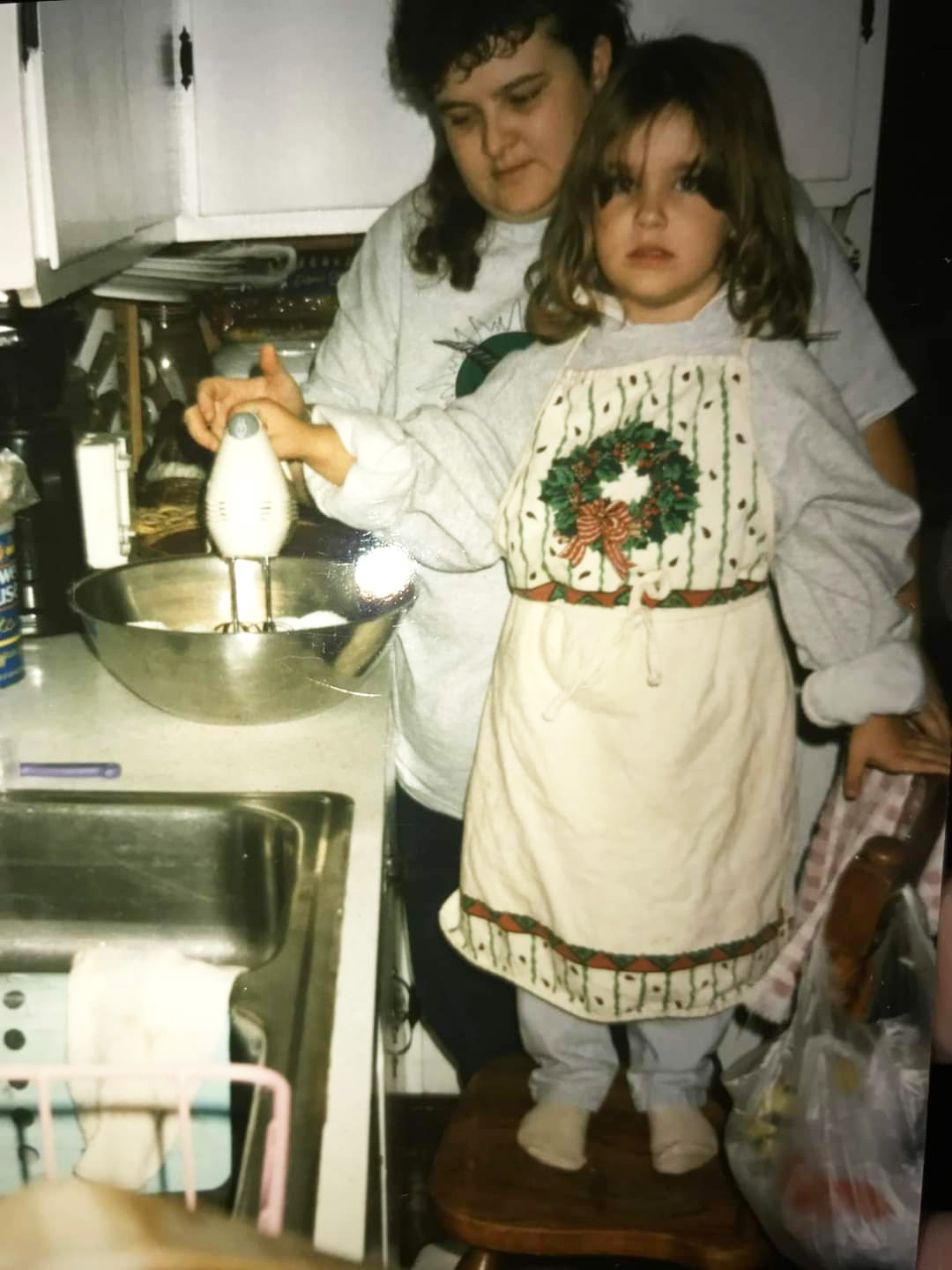 A child stands on a chair next to an adult woman in a kitchen